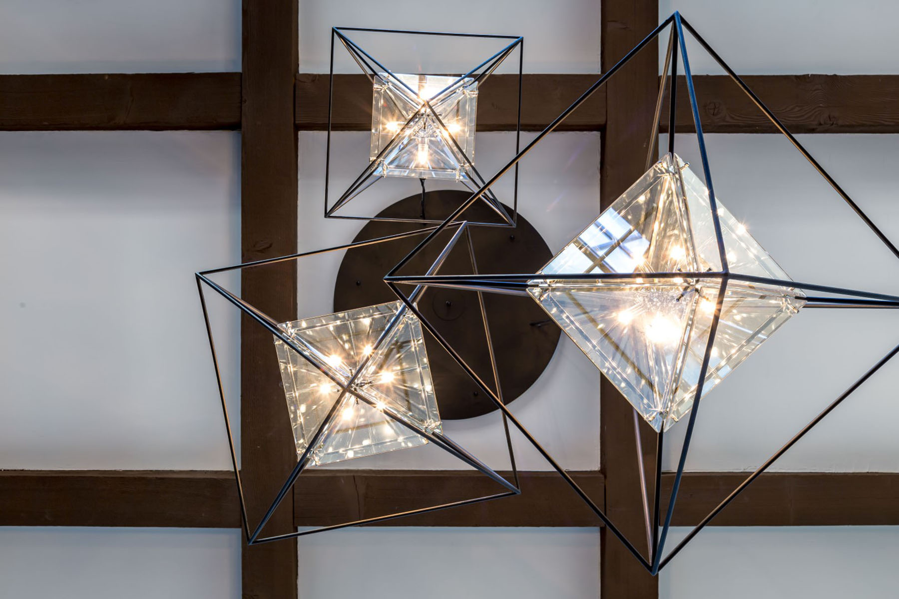 A striking custom chandelier designed by Trish Bowman Design, featuring glass and metal geometric shapes suspended beneath exposed wood ceiling beams.