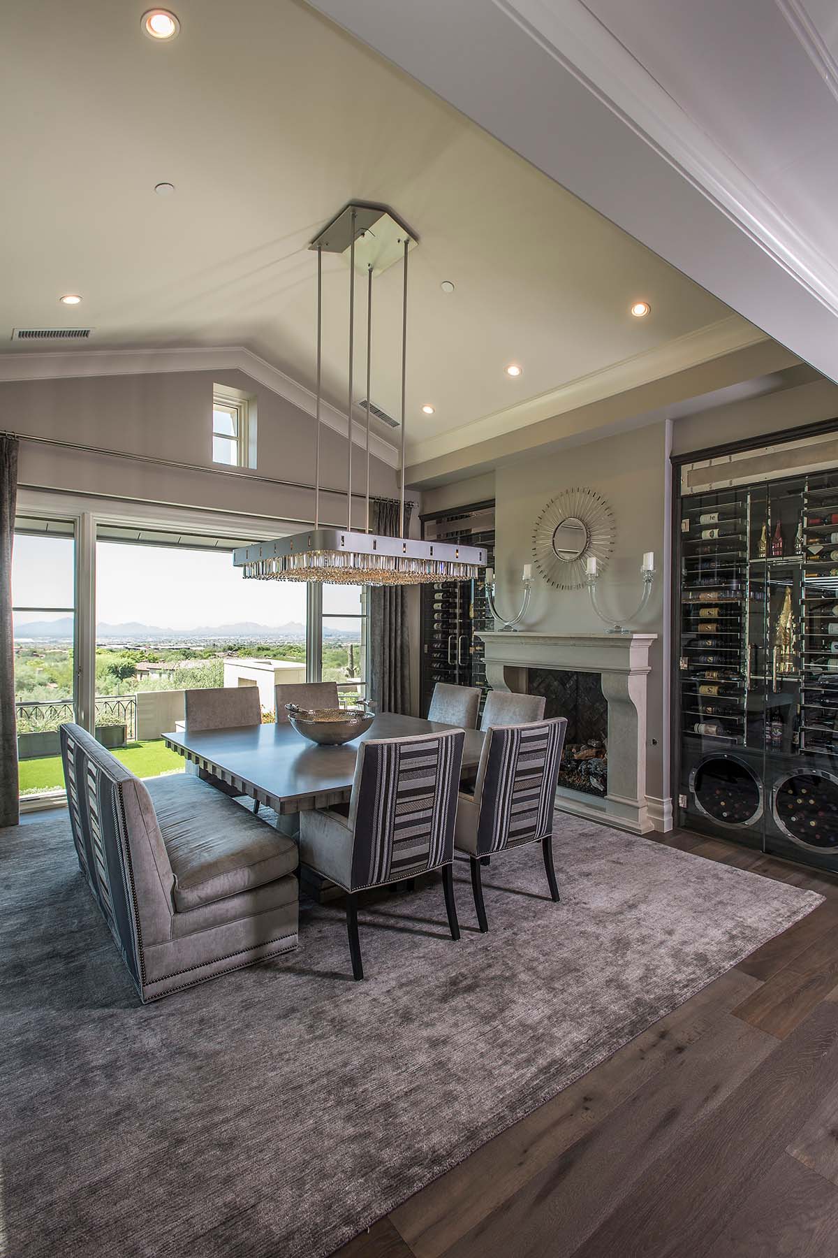Modern dining room design by Trish Bowman with vaulted ceilings, crystal chandelier, custom bench seating, and integrated wine storage overlooking scenic Arizona views.