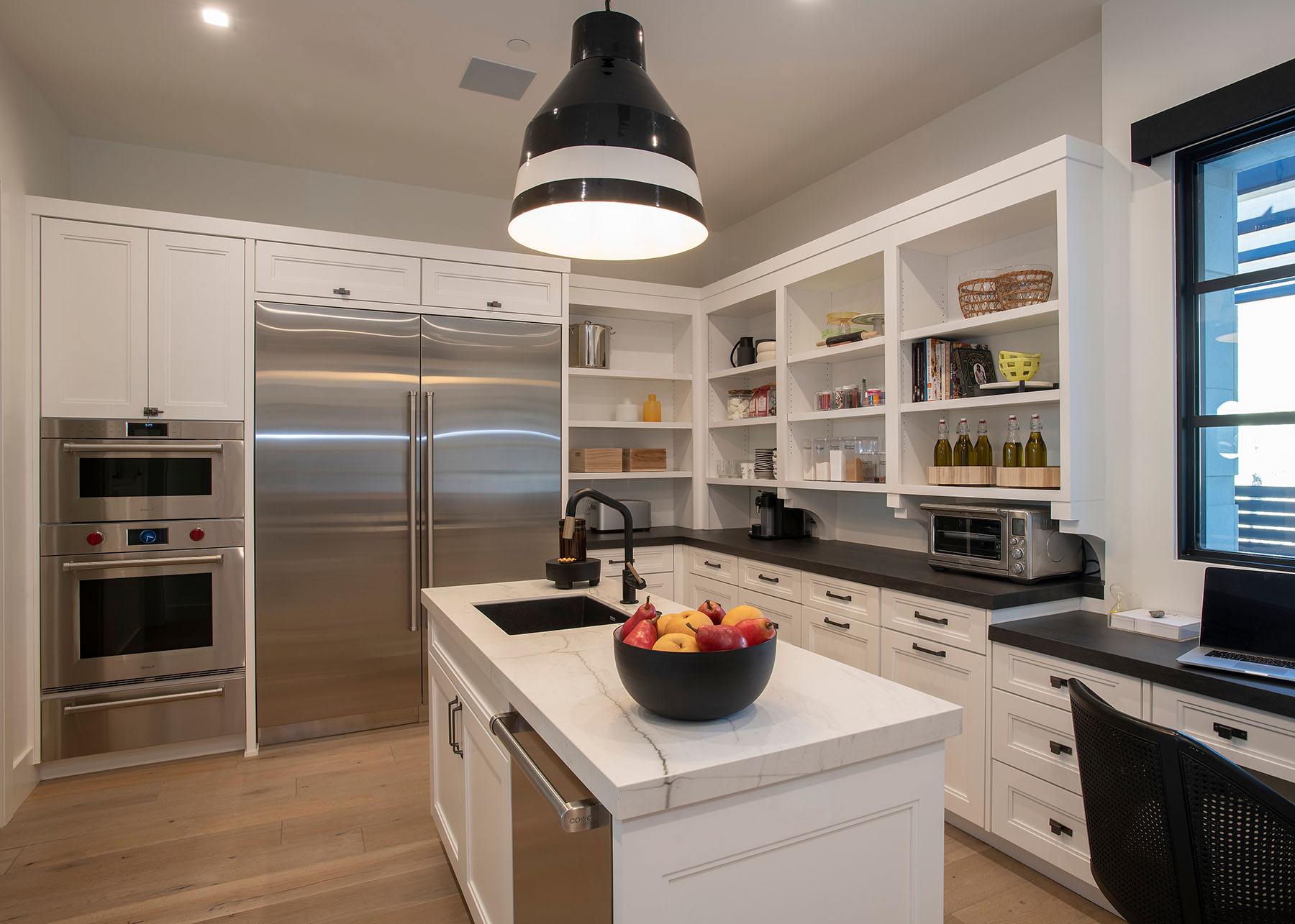 Custom kitchen design by Trish Bowman featuring white cabinetry, oversized pendant light, integrated appliances, and built-in desk with natural light.