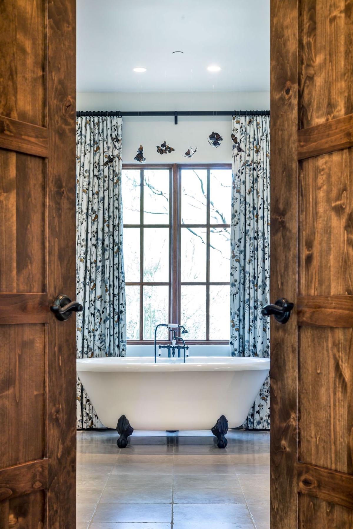 Freestanding clawfoot tub framed by floral drapes and butterfly decor, viewed through warm wood double doors.