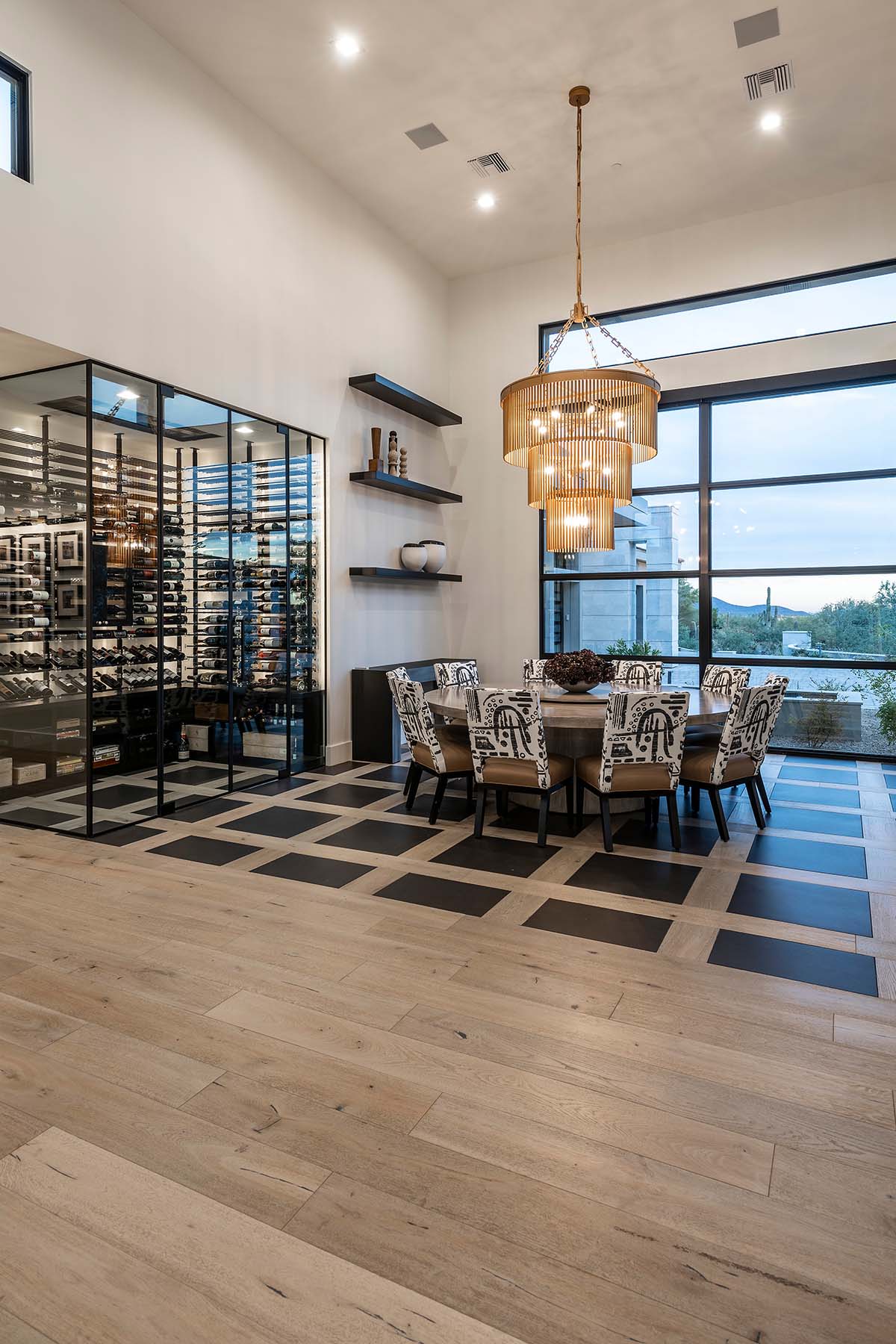 Statement dining room by Trish Bowman Design with tiered gold chandelier, custom patterned flooring, and glass-enclosed wine wall for refined entertaining.