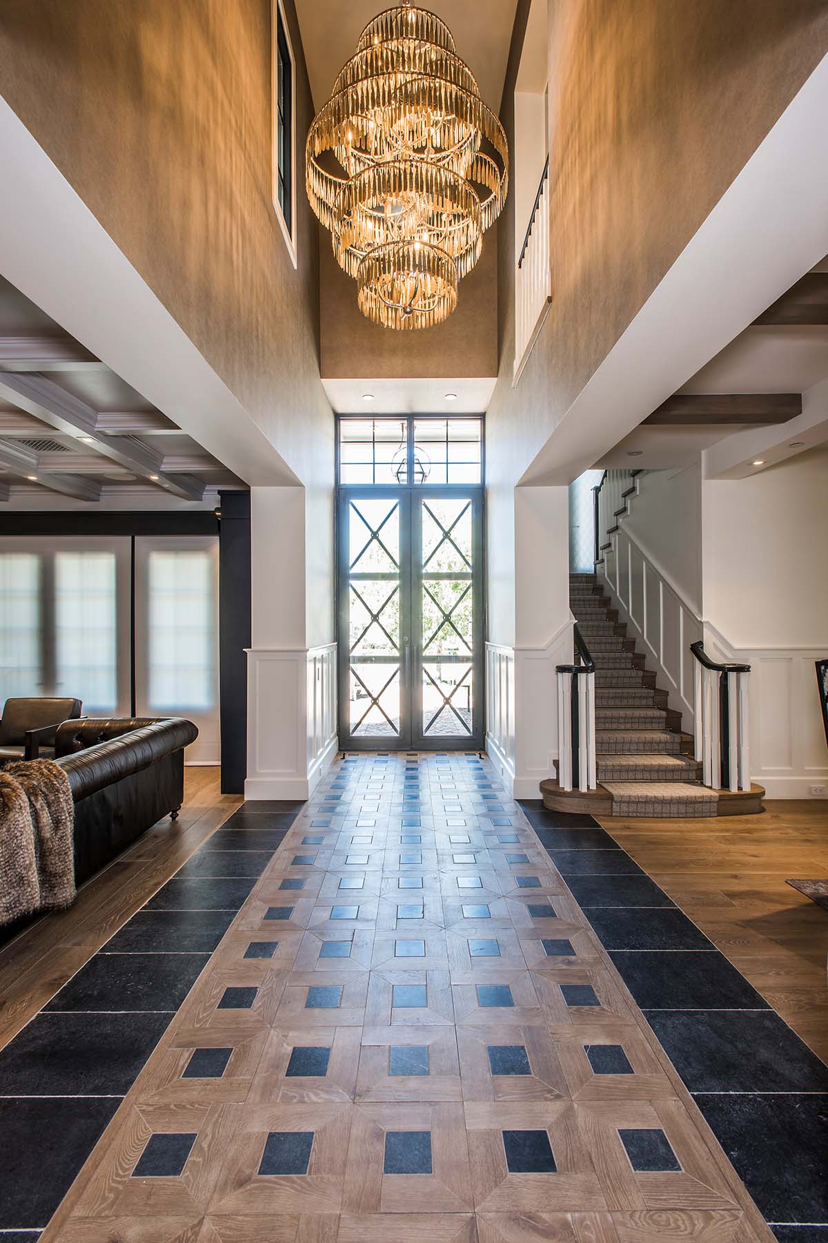 Grand entryway designed by Trish Bowman Design featuring dramatic chandelier, geometric inlaid flooring, and natural light pouring through custom glass doors.