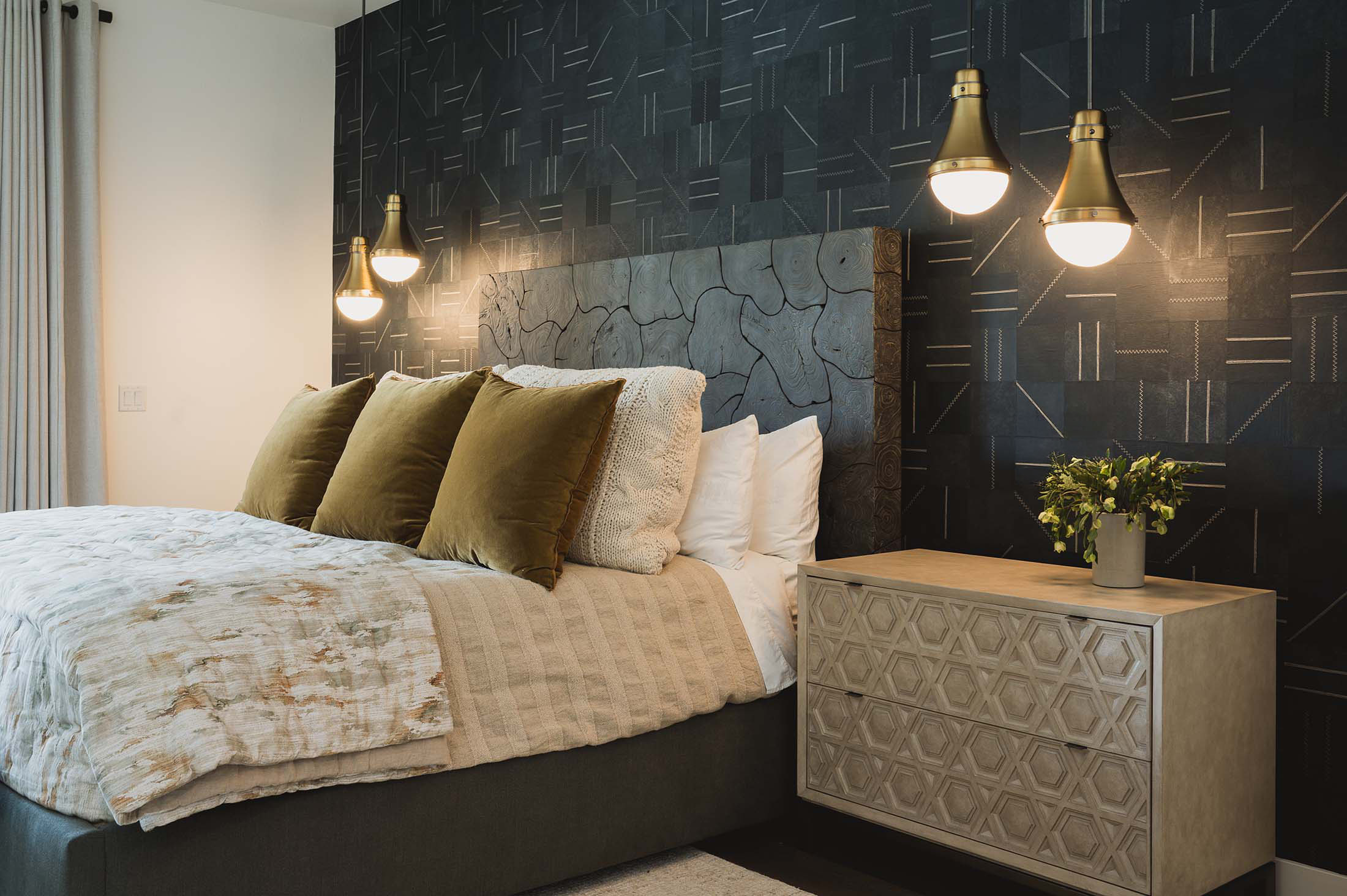 A wide angle of a modern bedroom with a textured black wall, gold lighting, a wood accent headboard, and patterned nightstands.